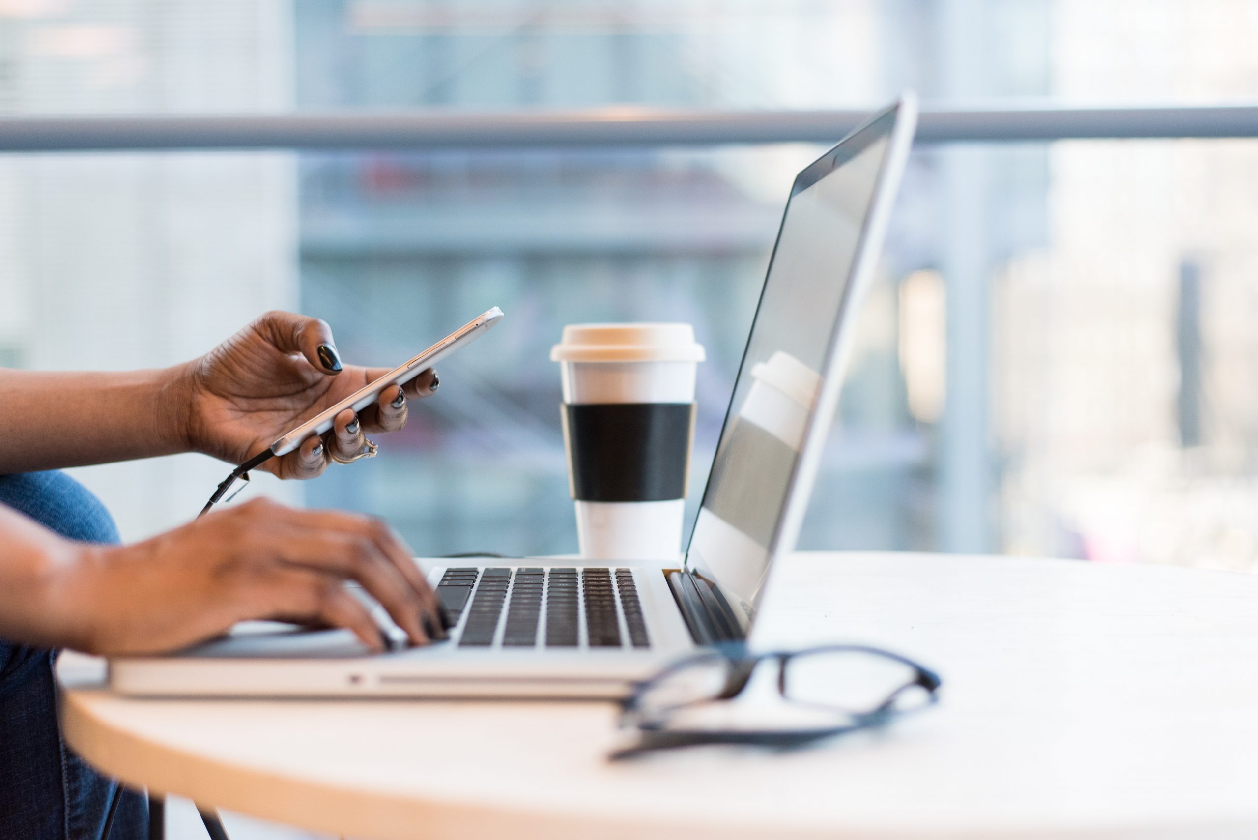 Image of brown hands with nail polish, holding a phone in one hand and typing on a laptop with the other. There is a coffee cup and a pair of glasses on the table with the laptop.